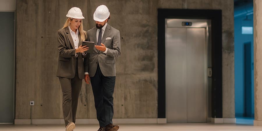 Two construction professionals in hard hats and business attire reviewing project details on a tablet in a hospital hallway near an elevator