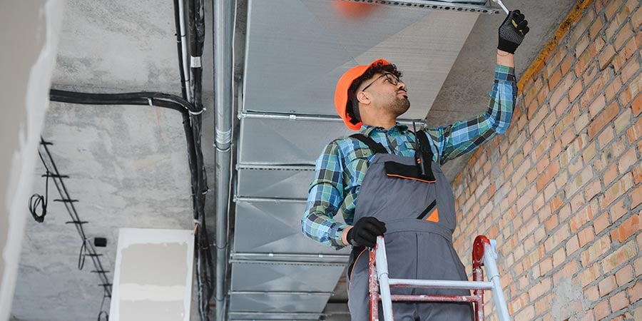 Construction worker on a ladder inspecting overhead ductwork above a ceiling, part of the containment system a negative air machine depends on