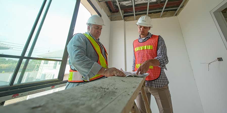 Two construction workers in hard hats and safety vests reviewing blueprints together inside an active healthcare construction site, with exposed ceiling framing and drywall visible around them.