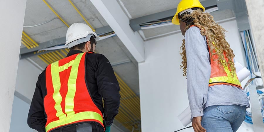 Two construction workers in hard hats and safety vests inspecting exposed ceiling wiring and structural beams inside an active building renovation.