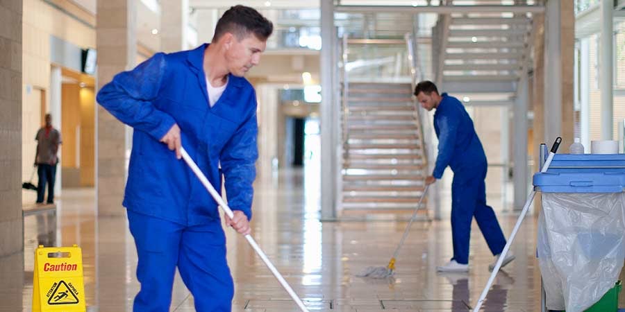 Two male cleaners in blue uniforms mopping the floor of a large atrium lobby, with a caution wet floor sign in the foreground.
