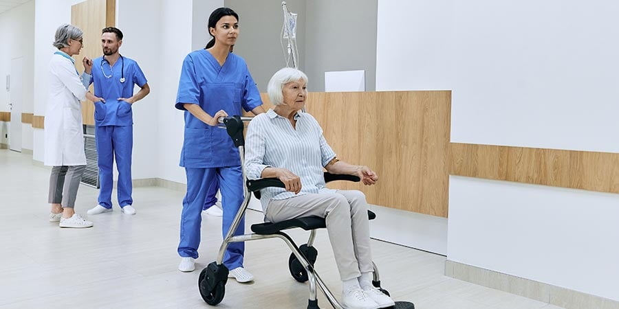 Nurse pushing an elderly patient in a wheelchair down a hospital hallway, with a doctor and another staff member talking in the background.