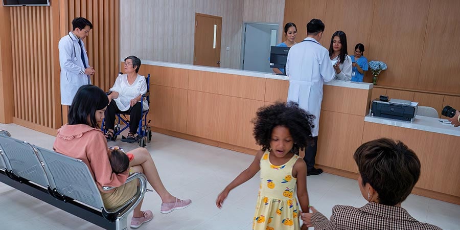 Healthcare facility waiting room with patients, family members, and clinical staff at a reception desk, illustrating a busy active facility environment