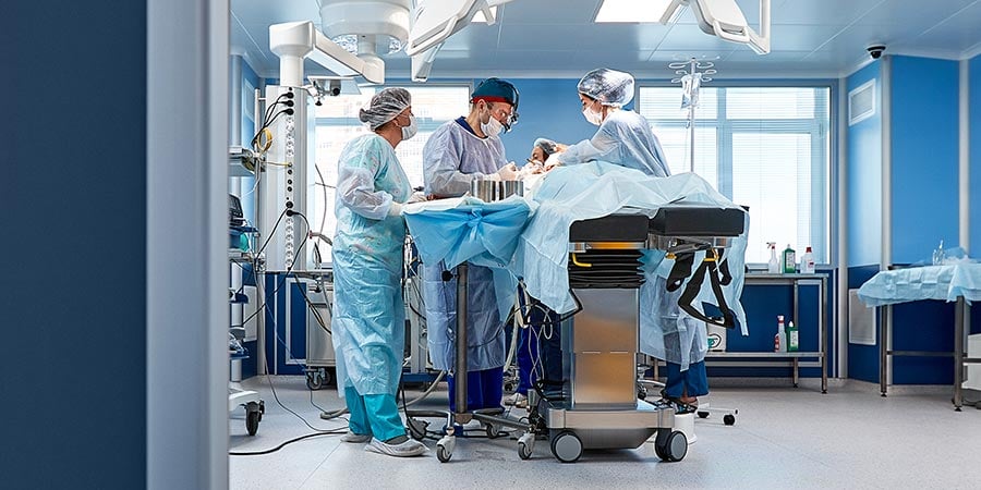 Surgical team in blue gowns and masks performing a procedure on a patient in a modern operating room, with monitoring equipment and a mobile filtration unit in the foreground.
