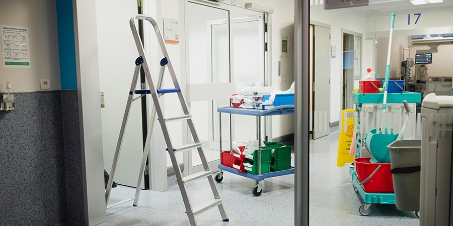 Cleaning cart and ladder positioned near an open hospital room door, illustrating routine maintenance work in an active care unit