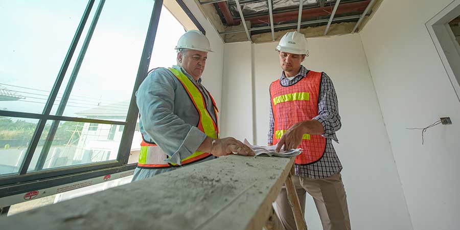 Two construction workers in hard hats and safety vests reviewing blueprints together inside an active healthcare construction site, with exposed ceiling framing and drywall visible around them.