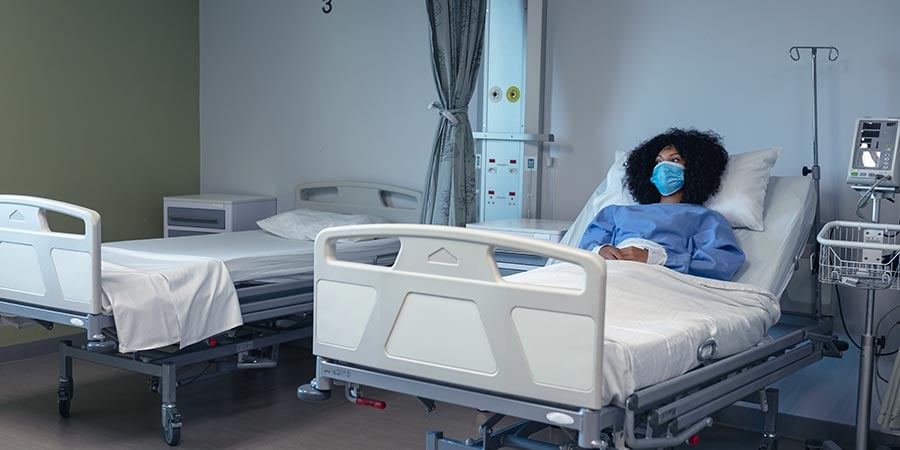Masked patient resting in a hospital bed beside an empty second bed, with a wall-mounted air filtration unit visible in the background.