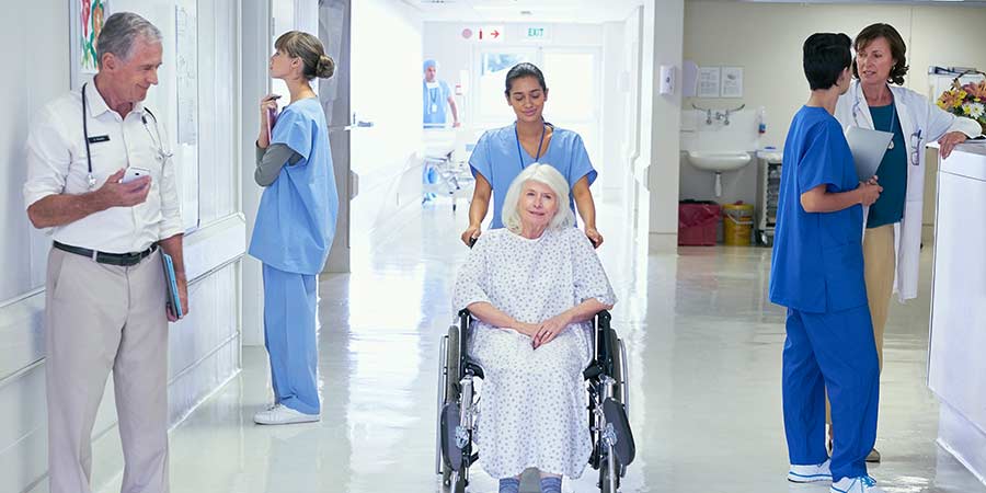 Healthcare staff transporting an elderly patient in a wheelchair down a hospital hallway with nurses and clinicians coordinating care in the background