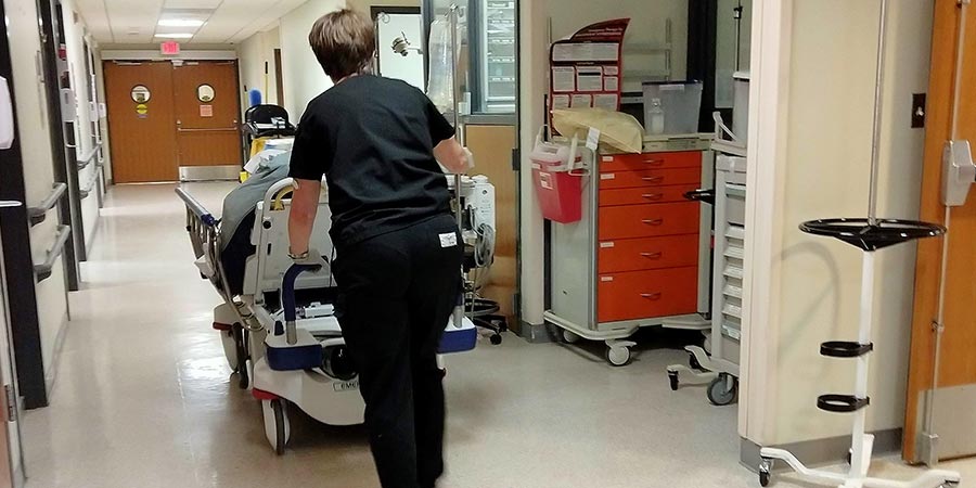 Healthcare staff member pushing a hospital stretcher down a corridor past a red supply cart and IV pole stand outside a patient care area.